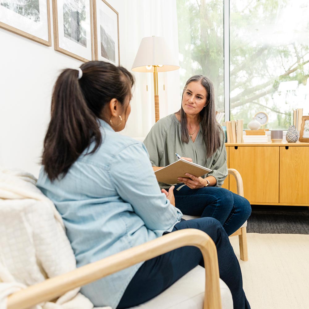 Two women sitting and talking in a bright, modern office — one taking notes while listening attentively to the other.