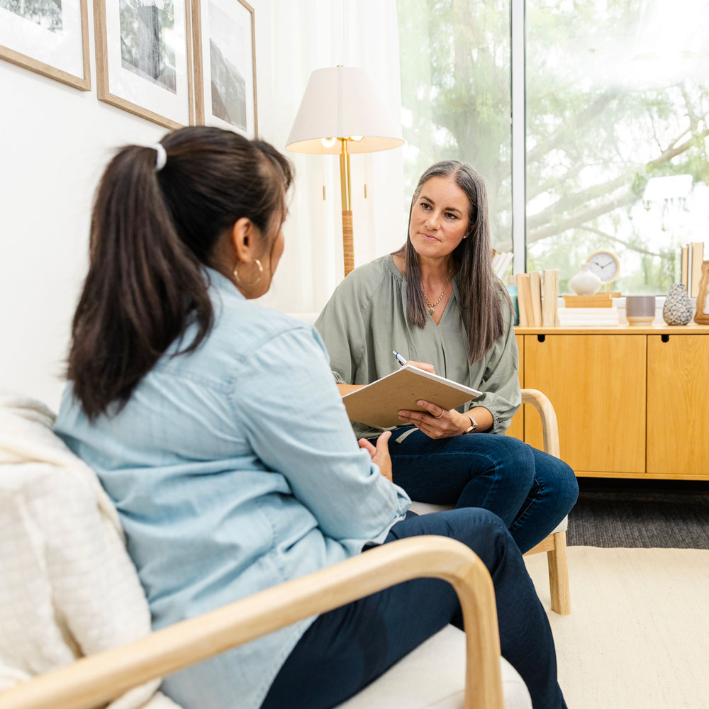 Two women sitting and talking in a bright, modern office — one taking notes while listening attentively to the other.