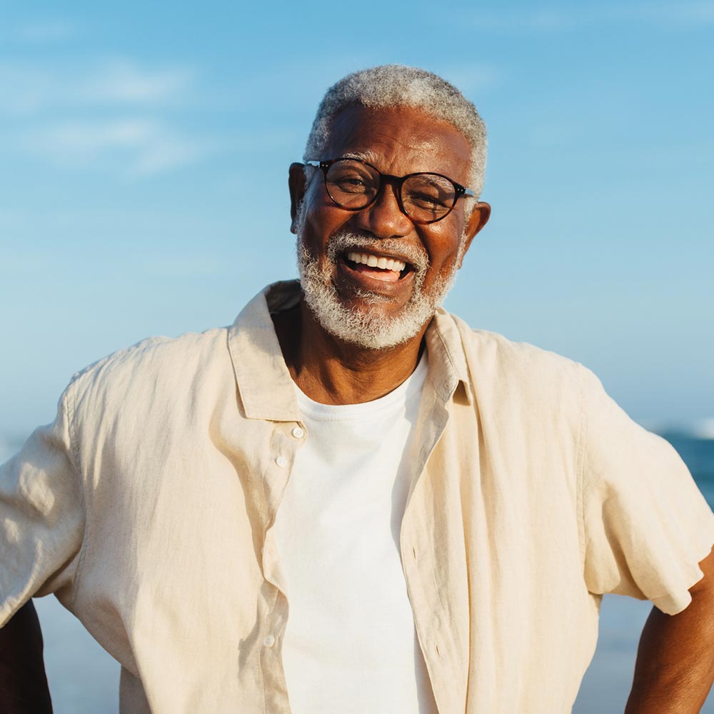 A smiling older man wearing glasses and a light shirt stands outdoors under a clear blue sky.