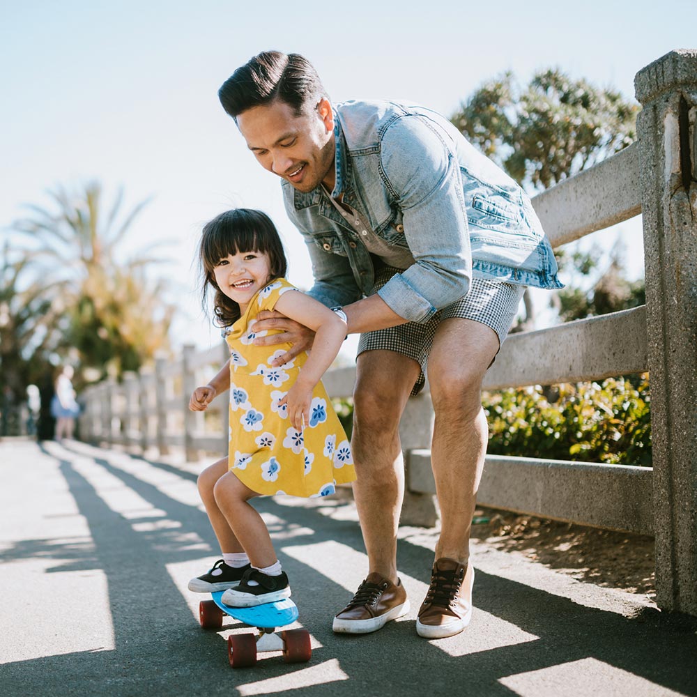 A smiling man helps a young girl ride a skateboard on a sunny day.