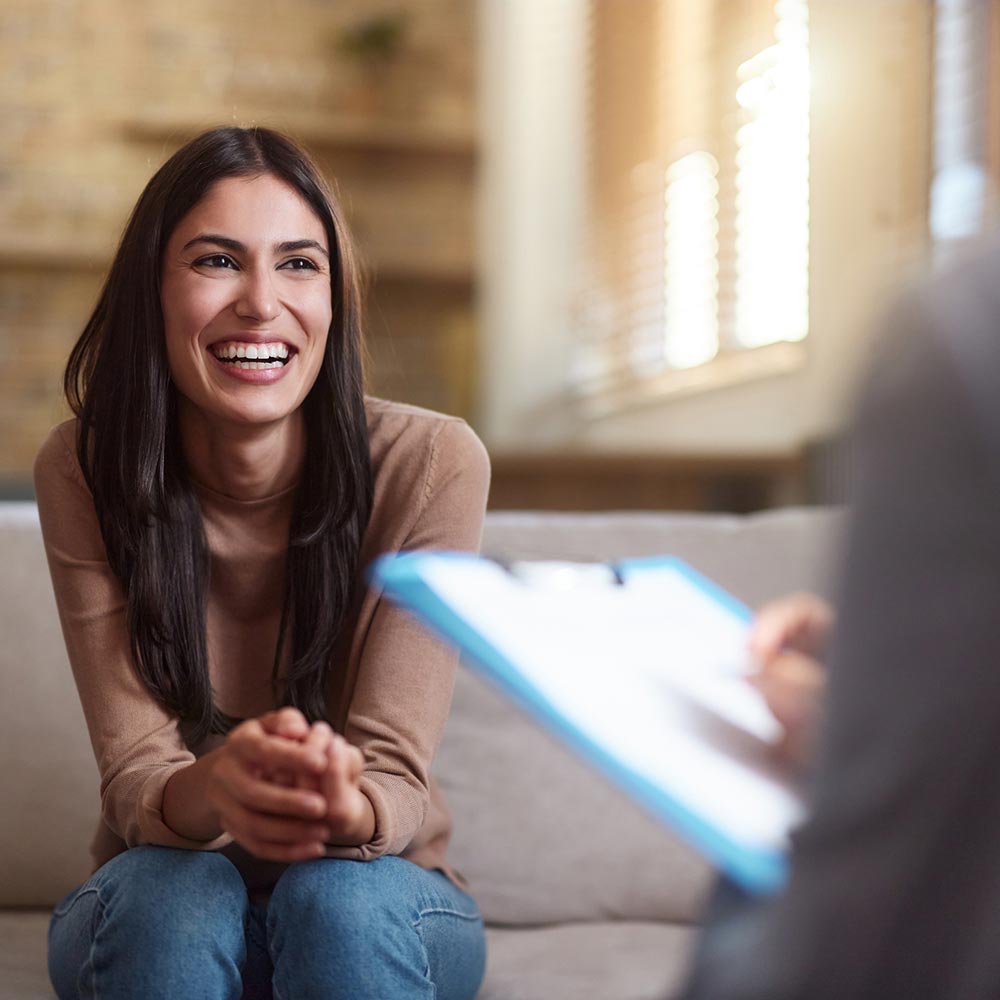 A woman smiles warmly while sitting on a couch during a conversation or therapy session.