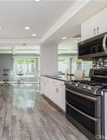 A modern kitchen with stainless steel appliances, white cabinets, and gray wood floors opening into a bright dining area.