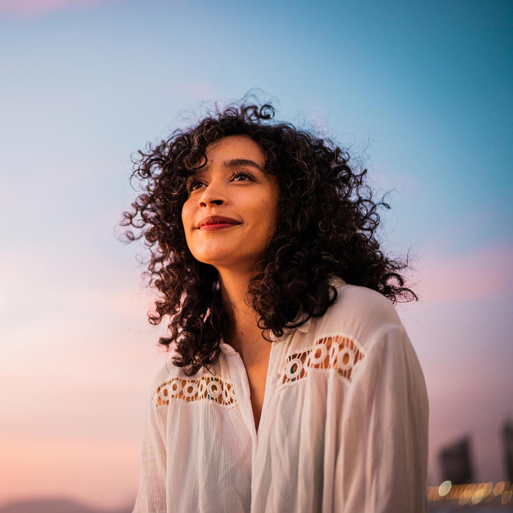 A woman with curly hair smiles softly while looking into the distance under a pastel sunset sky.