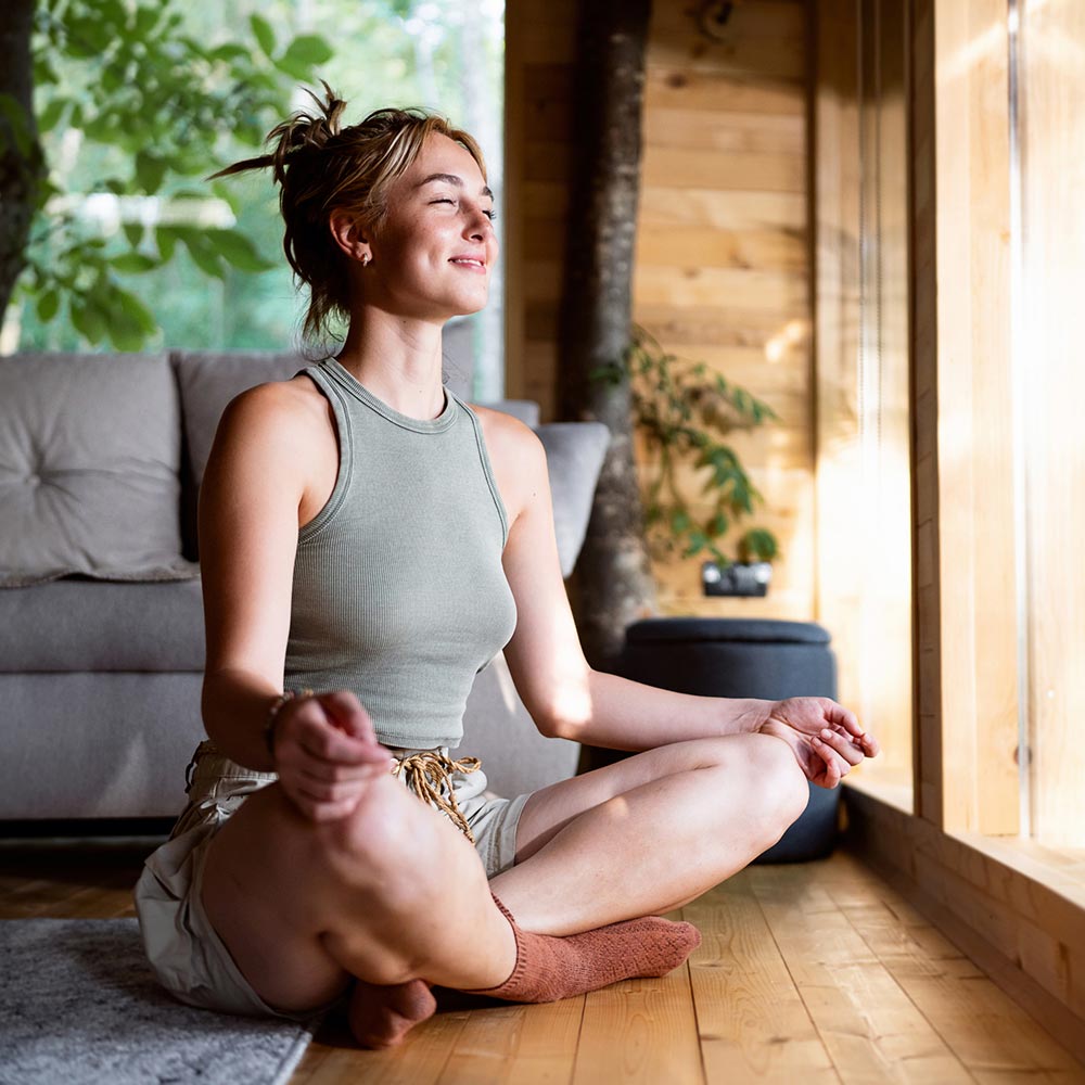 A woman sits cross-legged on the floor in a peaceful wooden room, meditating with a relaxed smile.