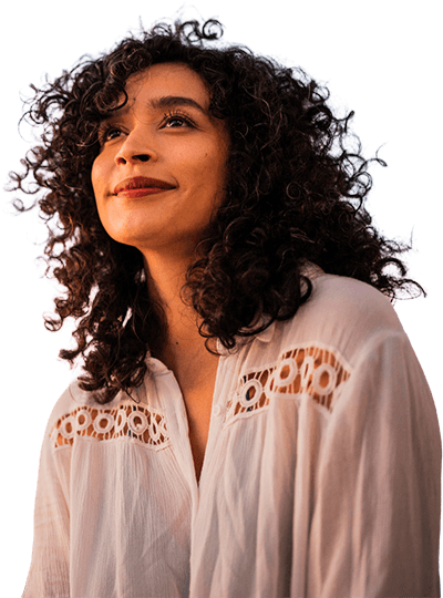 A woman with curly hair smiles softly while looking into the distance under a pastel sunset sky.